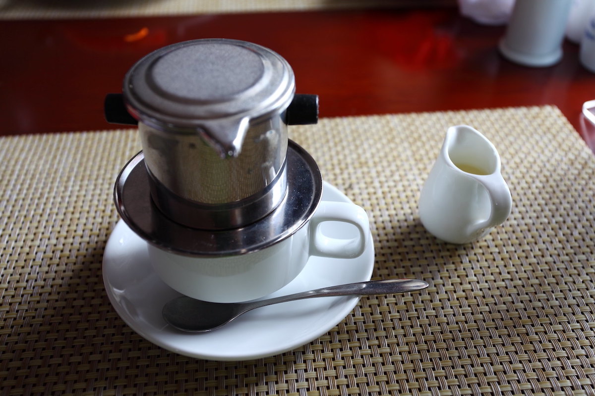 primer plano de una taza de café con leche y un platillo con un dispositivo de filtro de goteo de metal sobre la taza, haciendo café al estilo vietnamita