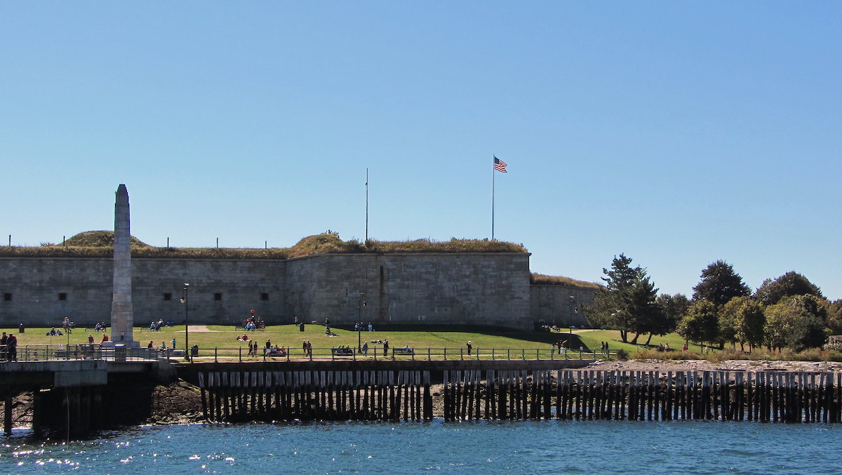 plano general de Castle Island Park en Boston. Un castillo de piedra en el fondo y gente haciendo un picnic en el césped frente a él