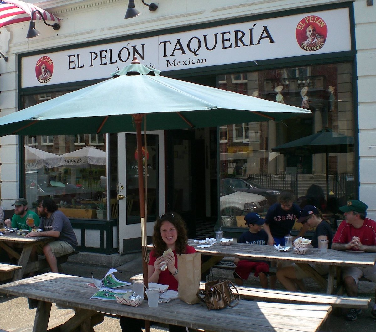 Una imagen del exterior de un restaurante en Boston con mesas de picnic y sombrillas verdes. Sobre la puerta, un letrero blanco con letras negras dice "EL PELON TAQUERIA"