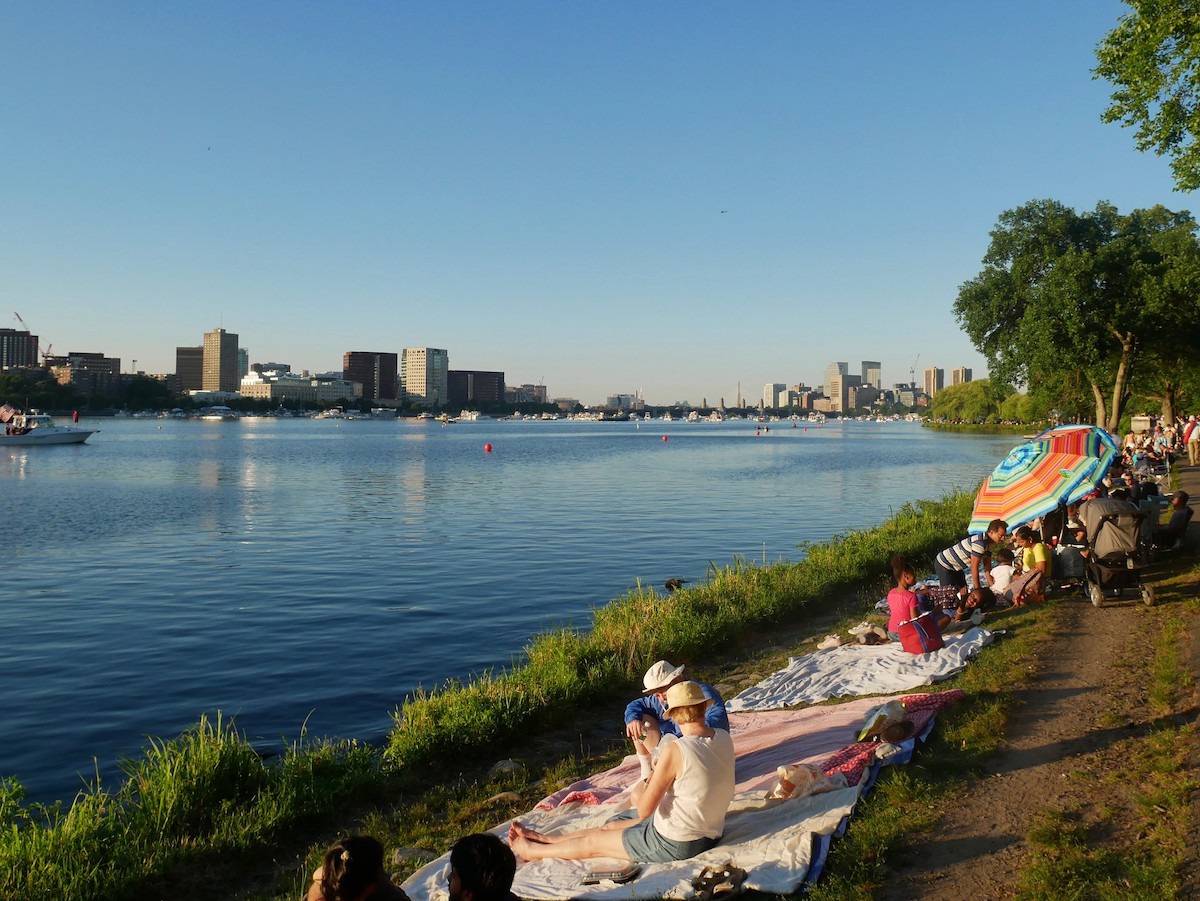 Hay muchas personas a orillas del río Charles sentadas sobre mantas y disfrutando de un excelente lugar para hacer un picnic en Boston el 4 de julio.