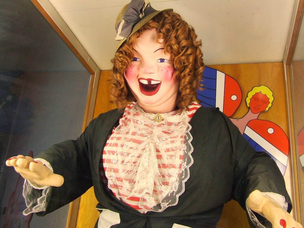 Primer plano de un personaje animatrónico llamado Laffing Sal. Está vestida con ropa de principios del siglo XX con un sombrero colocado a un lado de la cabeza. Ella tiene cabello rojo rizado, mejillas sonrosadas y una gran sonrisa con la boca abierta.