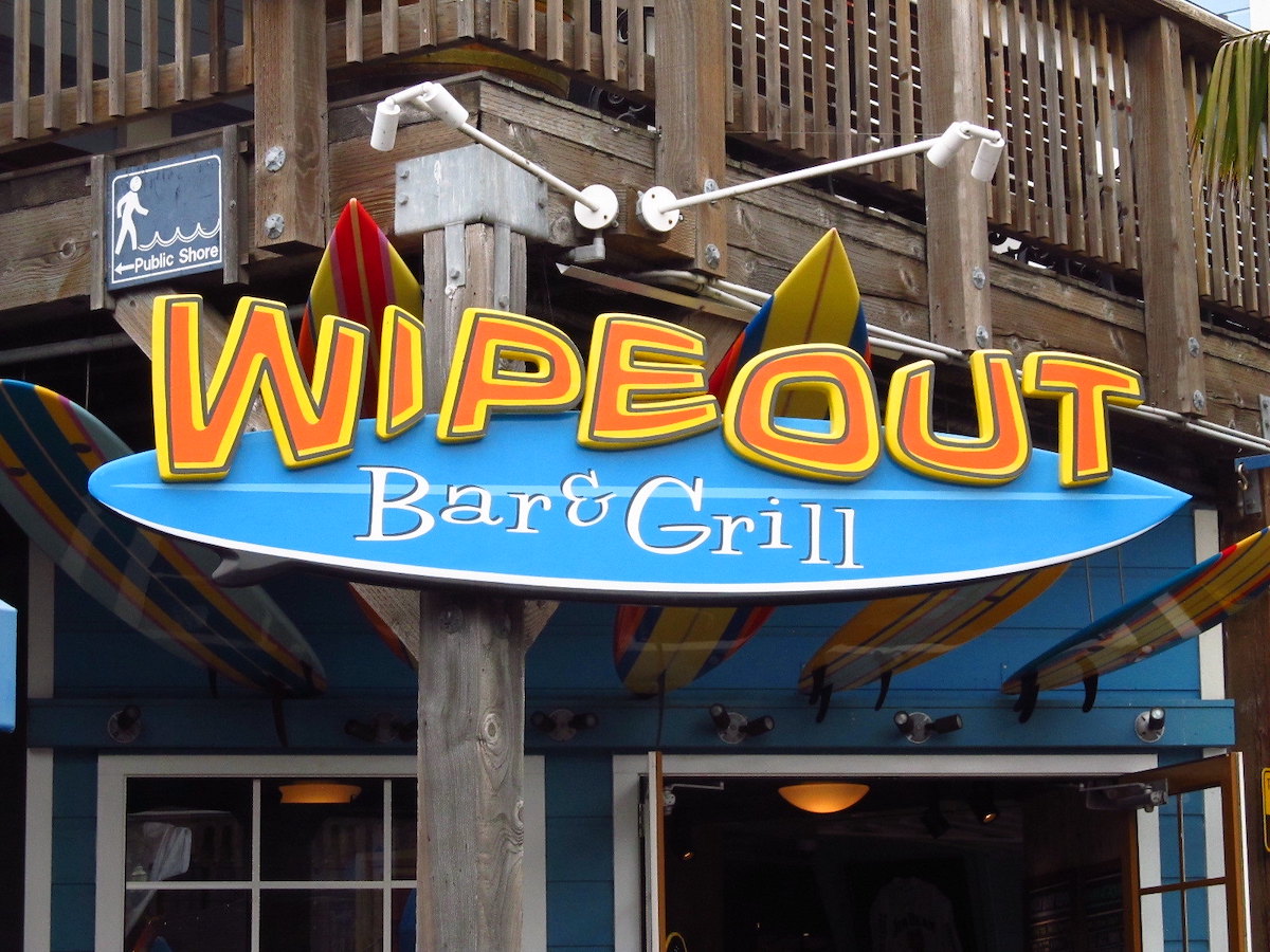 Foto del letrero de un restaurante que dice "Wipeout" en letras grandes de color naranja y amarillo sobre la forma de una tabla de surf azul brillante. Wipeout Bar & Grill es un restaurante en San Francisco Pier 39 .