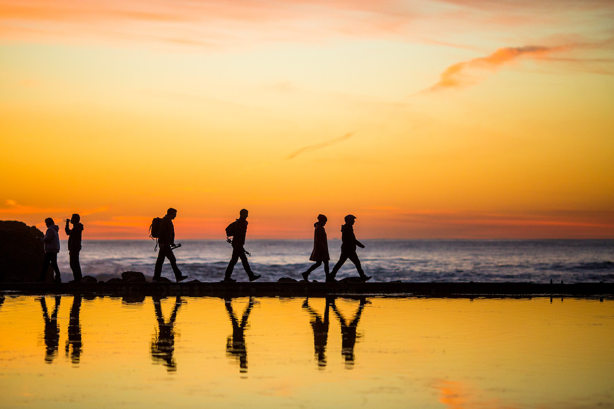 Cuatro siluetas se muestran caminando en el agua de los baños Sutro, el cielo naranja dorado al atardecer