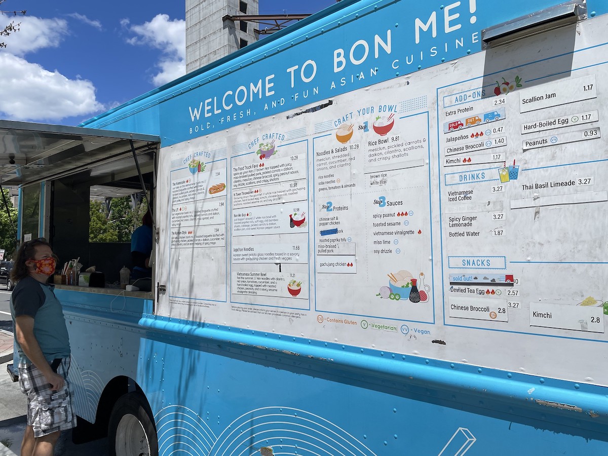 Una persona se para en la ventanilla de pedidos de un camión de comida azul en Boston. Un gran menú blanco con imágenes coloridas se extiende a lo largo del costado del camión. 