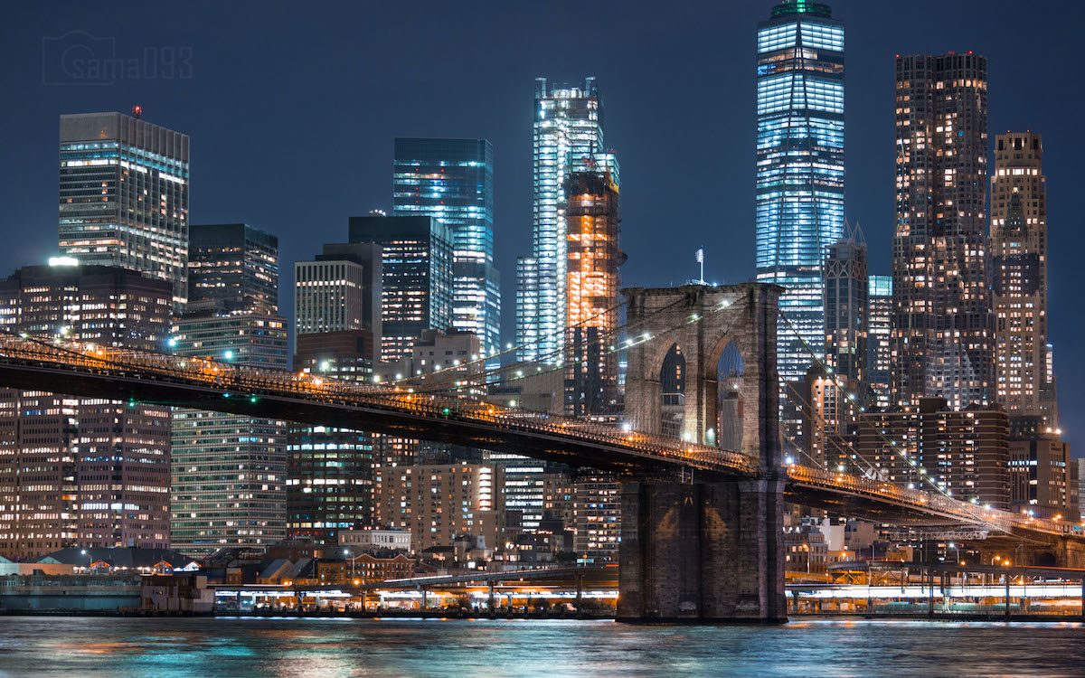 Una vista del horizonte de Manhattan y el puente de Brooklyn iluminados por la noche, tomada desde una masa de agua.