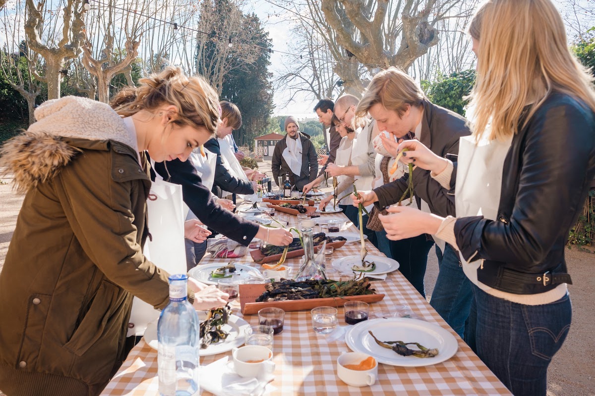 Un grupo de personas de pie alrededor de una mesa afuera comiendo verduras a la parrilla