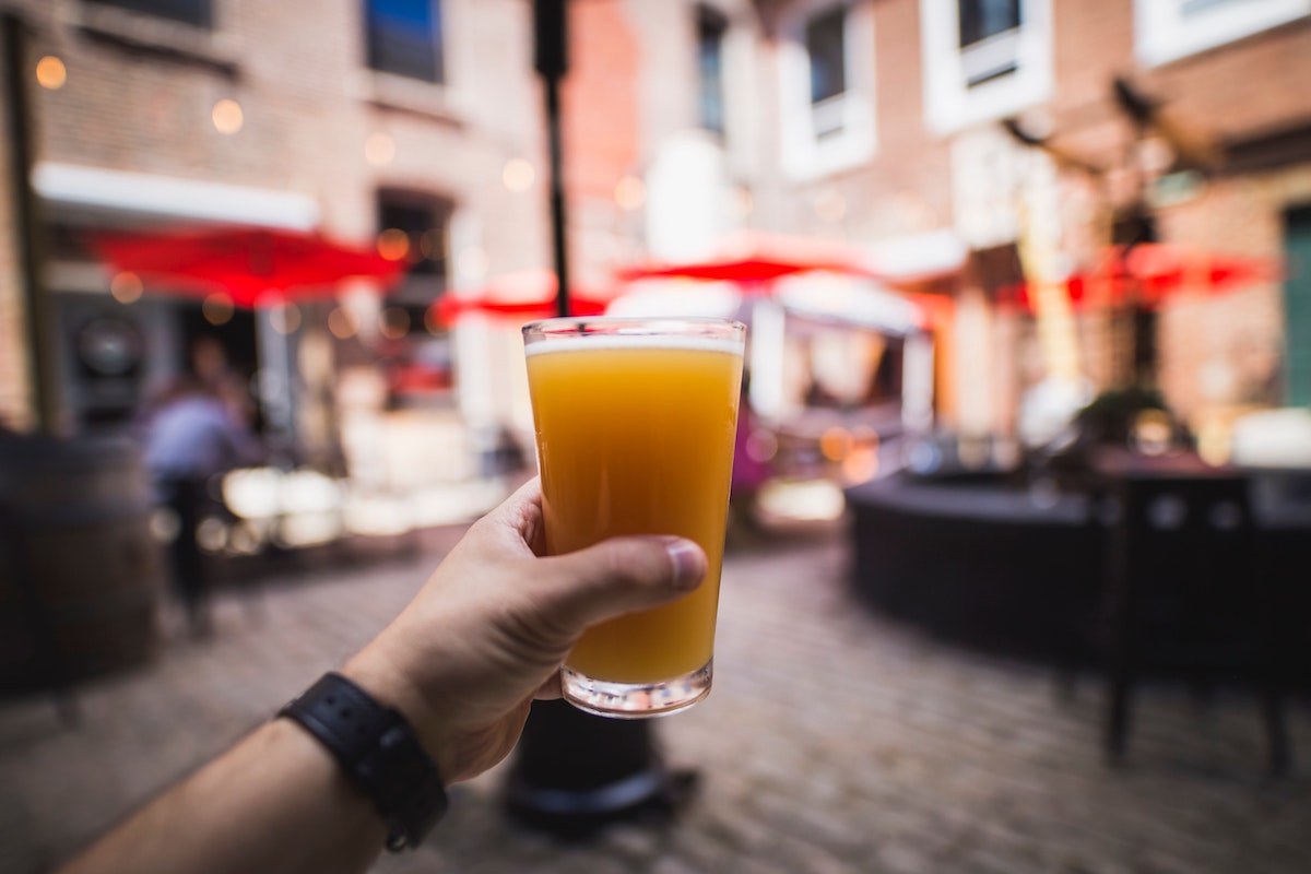 primer plano de la mano de una persona sosteniendo un vaso de cerveza y una plaza borrosa en el fondo