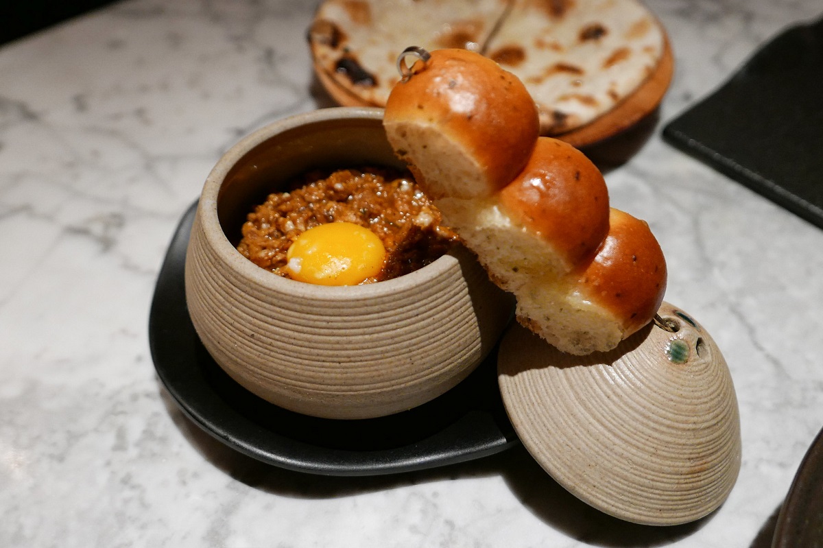 Un pequeño cuenco cilíndrico cubierto con comida india coronado con un huevo ensartado y bollos