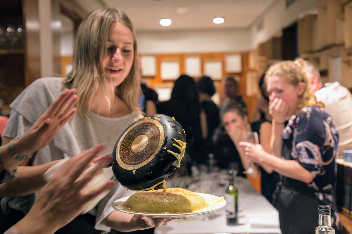 Una mujer sirviendo una tortilla española de una sartén a un plato mientras varias personas observan