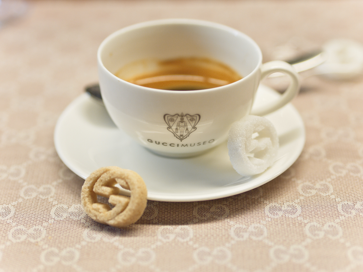 Una pequeña taza de espresso servida en una taza de porcelana con el emblema "Gucci Museo" con un terrón de azúcar blanco y uno moreno en forma de 2 G entrelazados