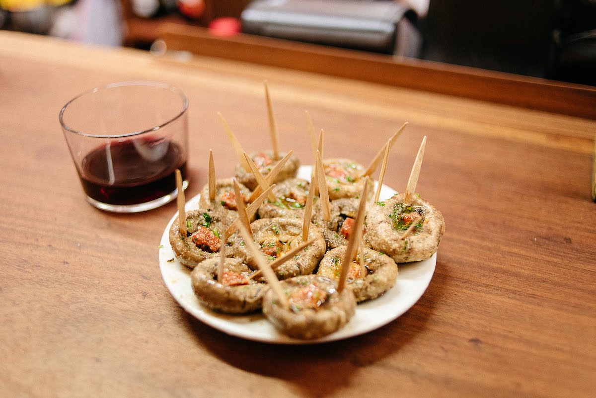 Un plato de champiñones a la plancha con chorizo junto a una copa corta de vino tinto.