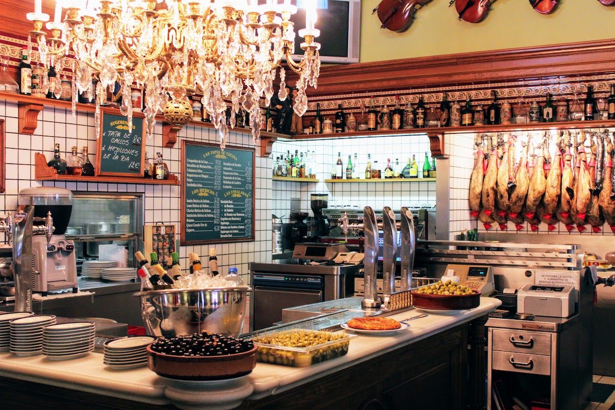 Interior de un típico bar español con grandes ollas de aceitunas en la parte superior de la barra y una gran lámpara de araña de cristal encima.